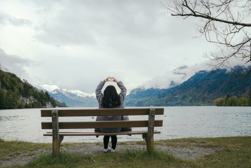 Woman sitting on bench in front of a lake, with her hands up forming a heart, looking joyful